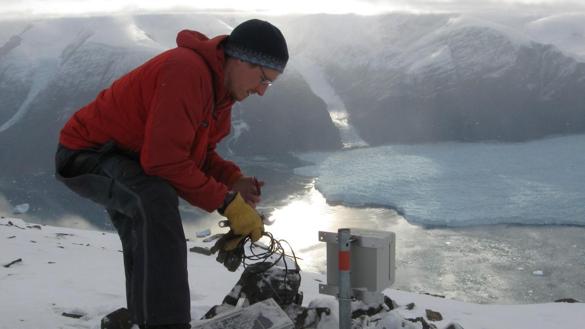Tim Bartholomaus leans over a seismometer with a glacier and mountain behind him.